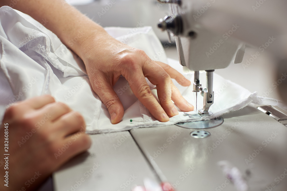 Close up side top view of cropped female hands sewing white fabric on professional manufacturing machine at seamstress workplace. Tailor hands holding textile for dress production. Blurred background