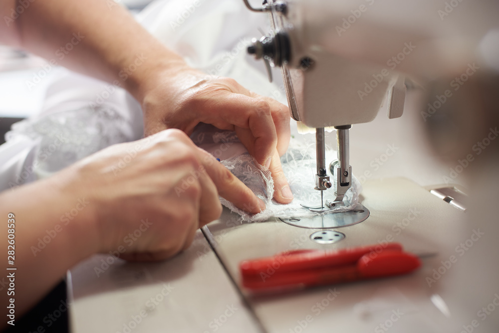 Female hands stitching white lace fabric on professional sewing machine at atelier. Seamstress hands pulling textile under presser foot. Scissors on background. Blurred background. Close up side view.