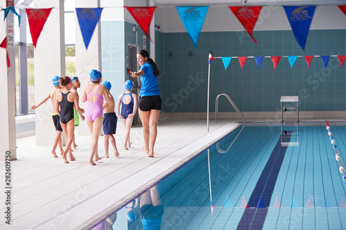 Female Coach Walks Children In Swimming Class Along Edge Of Indoor Pool