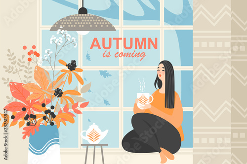 Cute girl sitting near the window and drinking tea on an autumn day. Autumn bouquet of red and orange leaves and dried flowers.