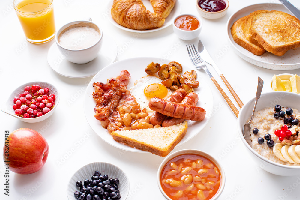 Top view flatlay with varieties of fresh breakfast: fried eggs with bacon and sausages, oatmeal with berries, fried toasts with jam and butter. White background.