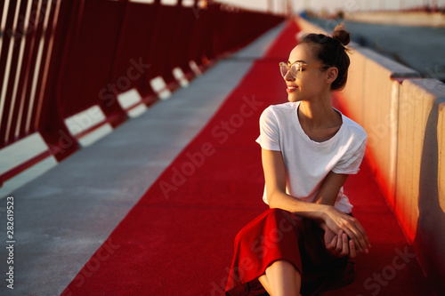 Bright summer lifestyle portrait of young pretty woman in eyewear, red skirt and white T-shirt, sitting on a bright red floor on the bridge.