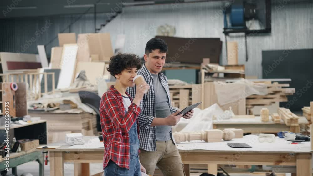 People walking in wooden furniture workshop talking discussing work holding coffee and tablet, girl is drinking to go drink in carton glass. Business and workplace concept. People walking in wooden furniture workshop talking discussing work holding coffee and tablet, girl is drinking to go drink in carton glass. Business and workplace concept.