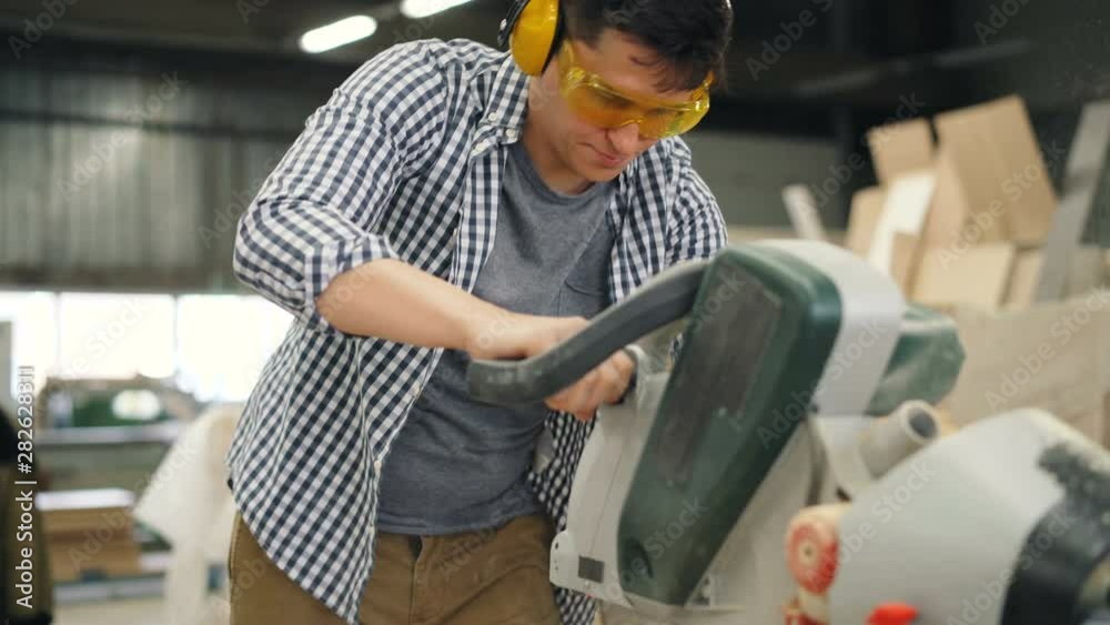 Carpenter young man in safety goggles and headphones is working with electric saw in workshop making handmade furniture. People and equipment concept. Carpenter young man in safety goggles and headphones is working with electric saw in workshop making handmade furniture. People and equipment concept.