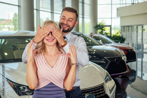 happy couple stand in front of new car, man covered wife eyes in showroom.surprised gift automobile