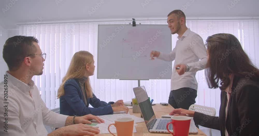Portrait of four collleagues meeting in the office indoors. Businessman drawing a graph on the whiteboard Portrait of four collleagues meeting in the office indoors. Businessman drawing a graph on the whiteboard