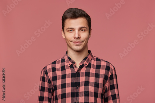 Portrait of young attractive man in checkered shirt, calmly looks at the camera, stands over pink background and smiles.