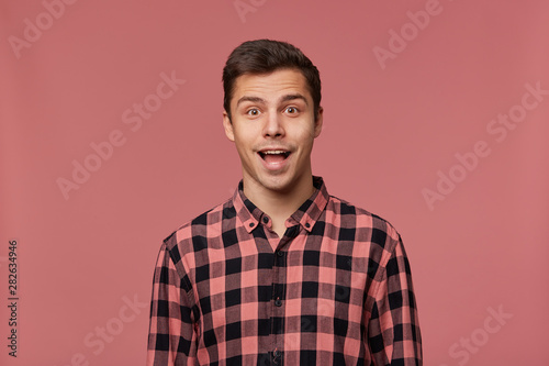 Portrait of young attractive happy amazed man in checkered shirt, looks at the camera with surprised expression, stands over pink background with wide open mouth.