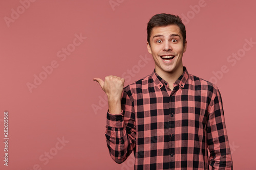 Young attractive cheerful man wears in checkered shirt, looks at the camera with happy expression, wants to turn your attention to copy space at the left side, stands over pink background.