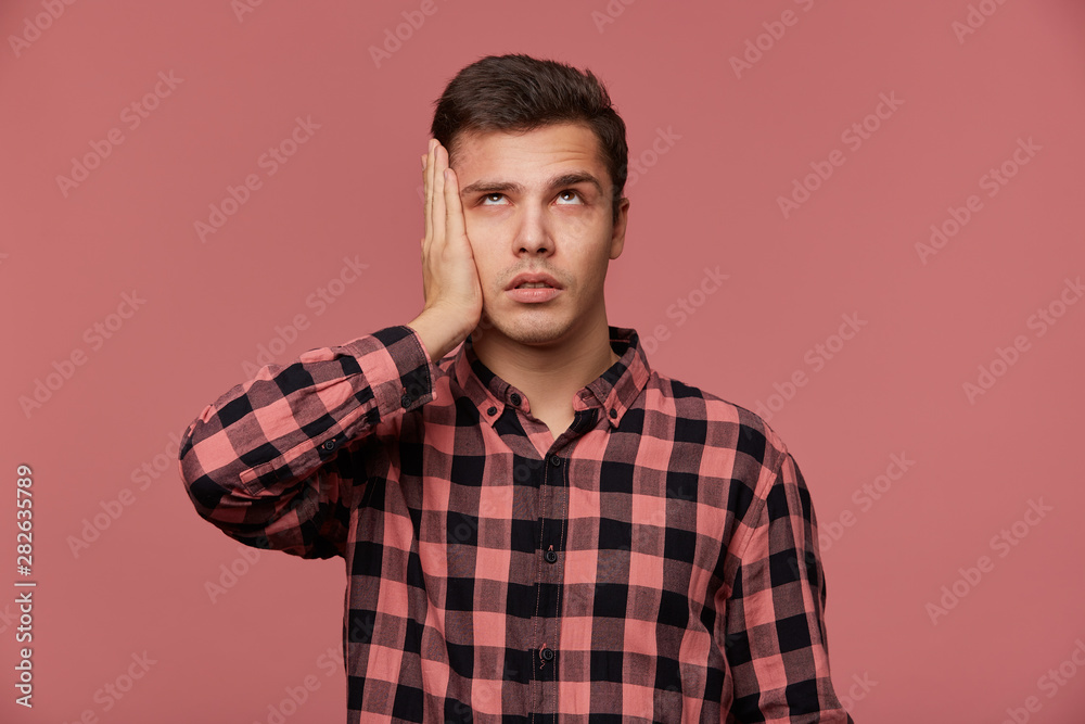 Portrait of young bored man in checkered shirt, looks up, stands over pink background and touches head, looks tired and sad. Portrait of young bored man in checkered shirt, looks up, stands over pink background and touches head, looks tired and sad.
