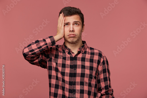 Portrait of young attractive man in checkered shirt, looks up, stands over pink background and touches head, looks tired and sad.