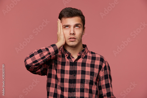Portrait of young bored man in checkered shirt, looks up, stands over pink background and touches head, looks tired and sad.