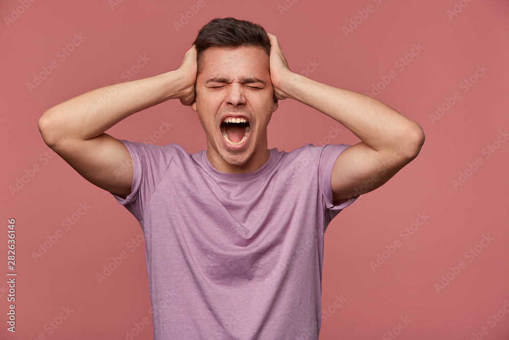 Portrait of young unhappy attractive man in blank t-shirt, stands over pink background and screaming, holds head, looks angry and unhappy. Portrait of young unhappy attractive man in blank t-shirt, stands over pink background and screaming, holds head, looks angry and unhappy.