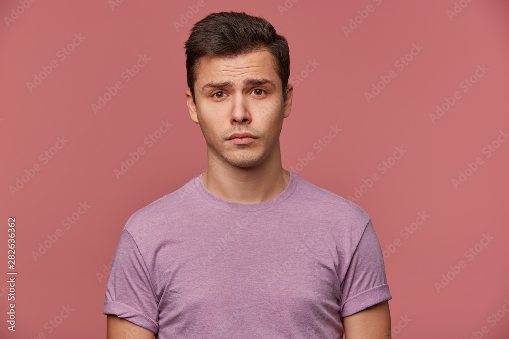 Close up of young offended attractive boy in blank t-shirt, stands over pink background and looks sad and unhappy.