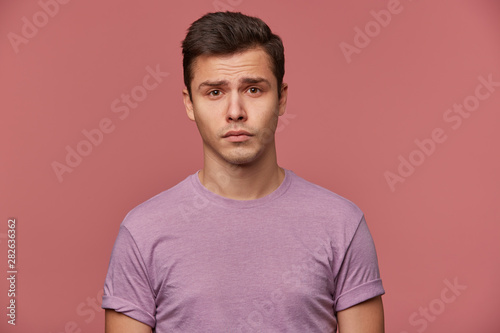Фотография Close up of young offended attractive boy in blank t-shirt, stands over pink background and looks sad and unhappy