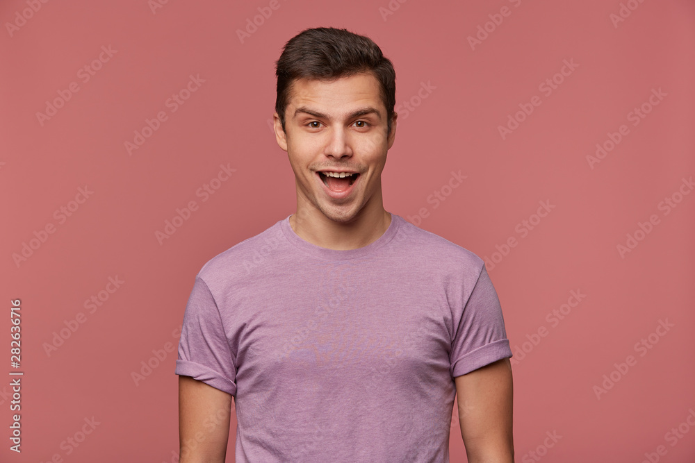 Obraz premium Portrait of cheerful handsome young man wears in blank t-shirt, looks at the camera with happy expression, stands over pink background.