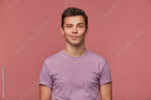 Portrait of handsome young man wears in blank t-shirt, looks at the camera with with a grin and happy expression, stands over pink background. © timtimphoto