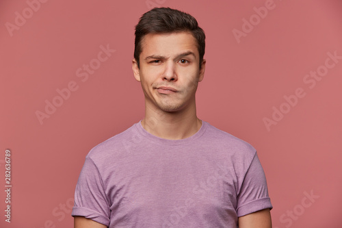 Portrait of handsome frowning young man wears in blank t-shirt, looks at the camera with with a grin and doubts, stands over pink background.