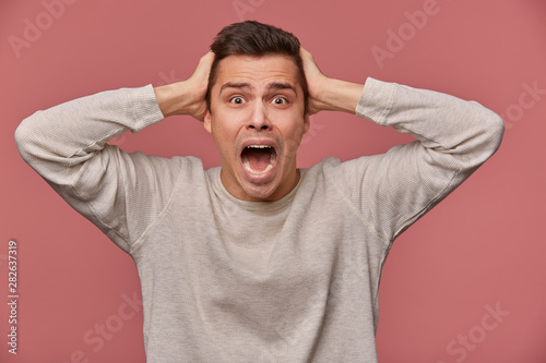 Stampa su tela Close up of young scared man in blank long sleeve, stands over pink background with closed eyes and screaming, looks afraid and unhappy, touches head