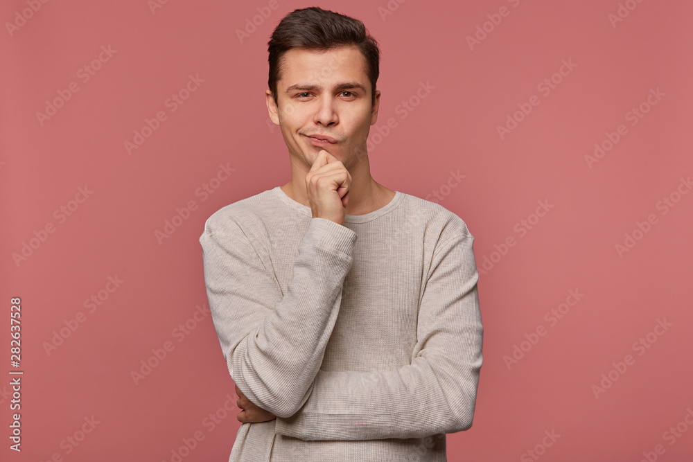 Young handsome thinking guy wears in checkered shirt, looks at the camera and touches chin, thinks about something, isolated over pink background. Young handsome thinking guy wears in checkered shirt, looks at the camera and touches chin, thinks about something, isolated over pink background.