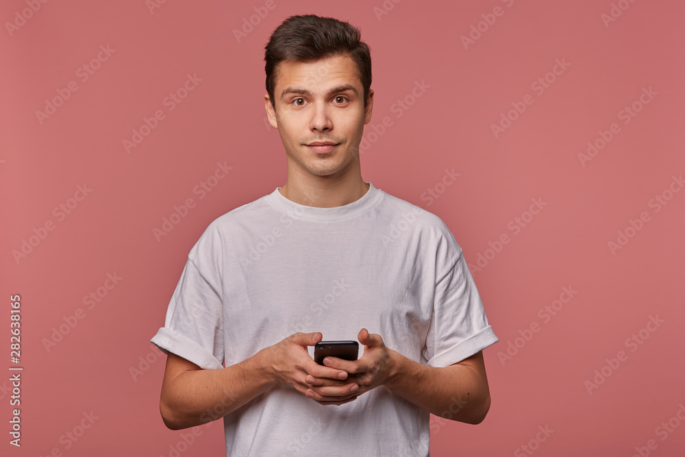 Portrait of young handsome cheerful guy wears in blank t-shirt, looks at the camera with calm expression, stands over pink background and chatting with friend on the telephone. Portrait of young handsome cheerful guy wears in blank t-shirt, looks at the camera with calm expression, stands over pink background and chatting with friend on the telephone.