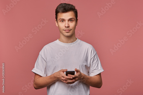 Portrait of young handsome cheerful guy wears in blank t-shirt, looks at the camera with calm expression, stands over pink background and chatting with friend on the telephone. © timtimphoto