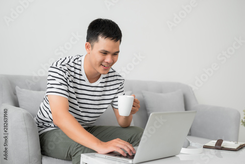 Young man concentrating on laptop computer screen, sitting at desk in living room, holding mug.?