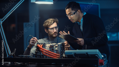 Two Professional Automotive Engineers with a Tablet Computer and Inspection Tools are Having a Conversation While Testing an Electric Engine in a High Tech Laboratory with a Concept Car Chassis.