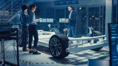 Automotive Design Engineers Talking while Working on Electric Car Chassis Prototype. In Innovation Laboratory Facility Concept Vehicle Frame Includes Wheels, Suspension, Engine and Battery. © Gorodenkoff