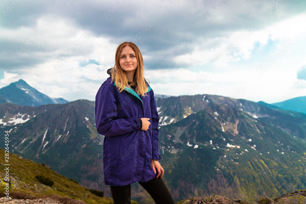 Naklejka premium Tourist adventures. Blonde girl in a warm gray sweater climbed the peak of the mountain and looks at the photographer camera.