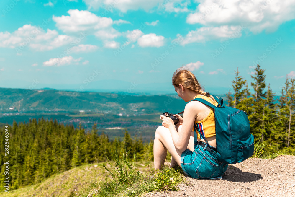 blonde cute girl with a backpack and glasses sits on a mountain and enjoys the beautiful hills of the mountains on a sunny day.