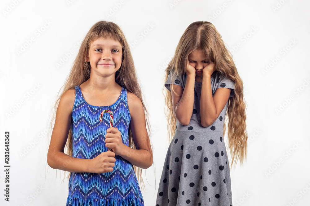 Girl joyfully holds candy, sad other girl in the background Girl joyfully holds candy, sad other girl in the background