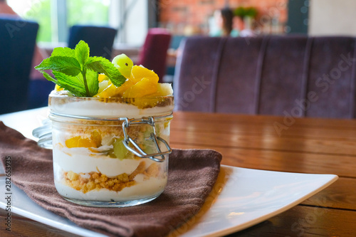Healthy layered fruit dessert in glass jar. Kiwi, mango, pineapple, peach, chia seeds, mascarpone, whipped cream, mint and granola on wooden table. Blurred restaurant on background. Sweet menu concept