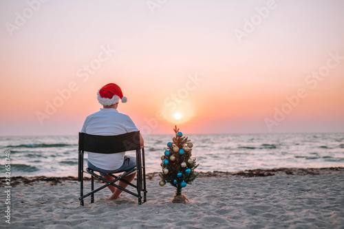 A guy in a santa hat on the beach with a Christmas tree at sunset.