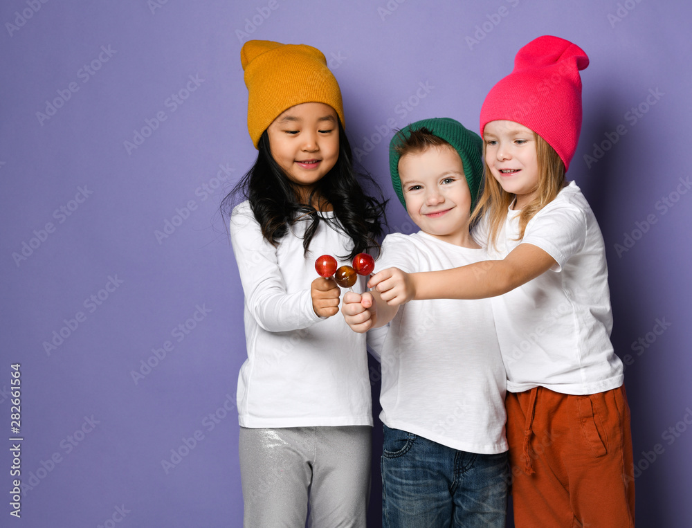 Joyful playing friends kids in white t-shirts and colorful hats hold close together thier lollipops and smiles Joyful playing friends kids in white t-shirts and colorful hats hold close together thier lollipops and smiles