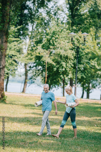 cheerful mature man and woman walking together in park while holding yoga mats