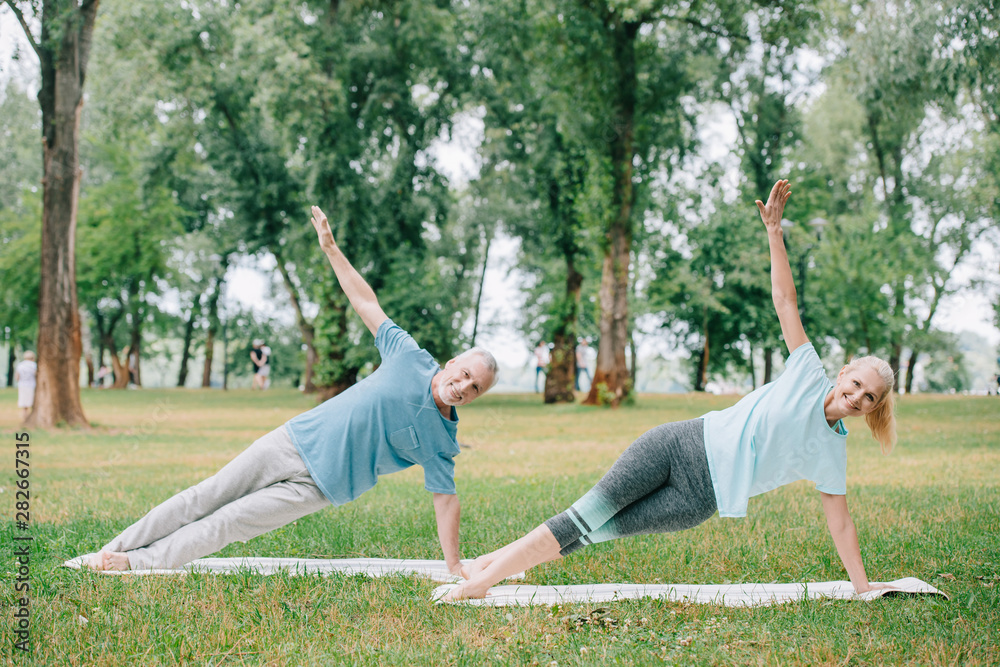 positive, mature man and woman doing side plank exercise while practicing yoga on lawn in park positive, mature man and woman doing side plank exercise while practicing yoga on lawn in park
