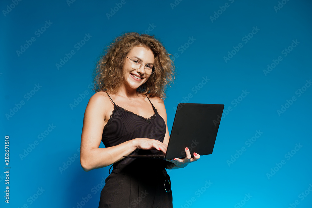 Portrait of happy young woman holding laptop computer over blue background. Brunette curvy hair and clever glasses.