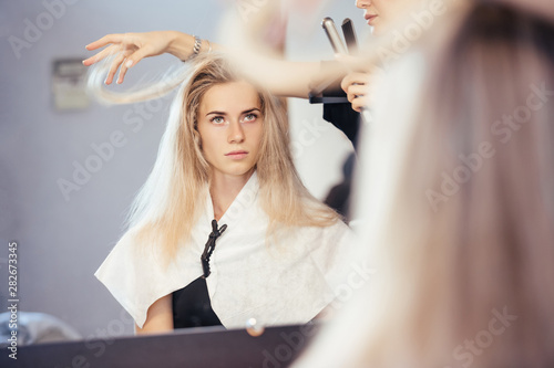 Side view of a beautiful young blonde girl looking at herself in the mirror while creating a beautiful image of makeup and hairstyle in a beauty salon with a master. The concept of a new image