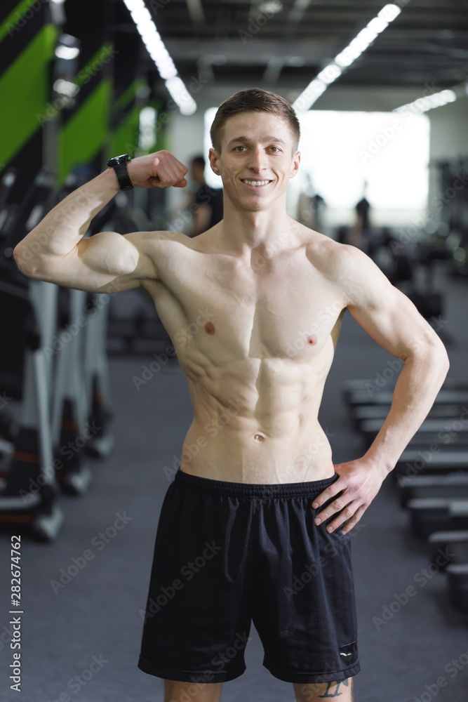 Obraz premium Portrait of sportsman with perfect body standing at the gym after workout and showing his biceps