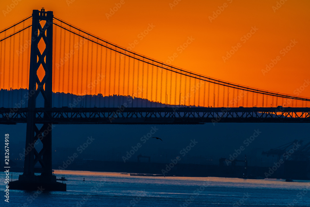 Fototapeta premium Beautiful summer morning sunrise behind mountain in California gives silhouette to the iconic Oakland Bay Bridge in San Francisco over the ocean.Famous travel location landmark in the west coast city.