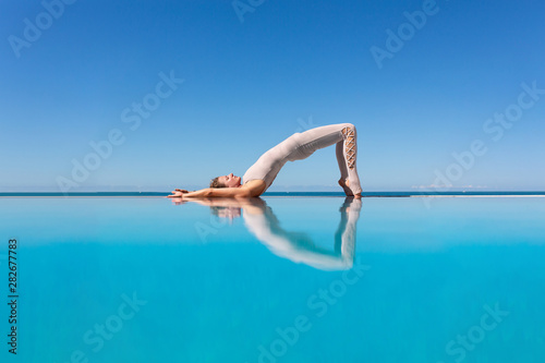 Beautiful athletic slim woman doing Ardha Chakrasana standing on the edge of a blue water pool against the sky while relaxing at a spa resort. Copyspace © Rithor