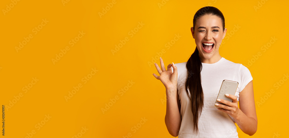Woman Holding Phone And Gesturing OK Sign Over Yellow Background - obrazy, fototapety, plakaty 