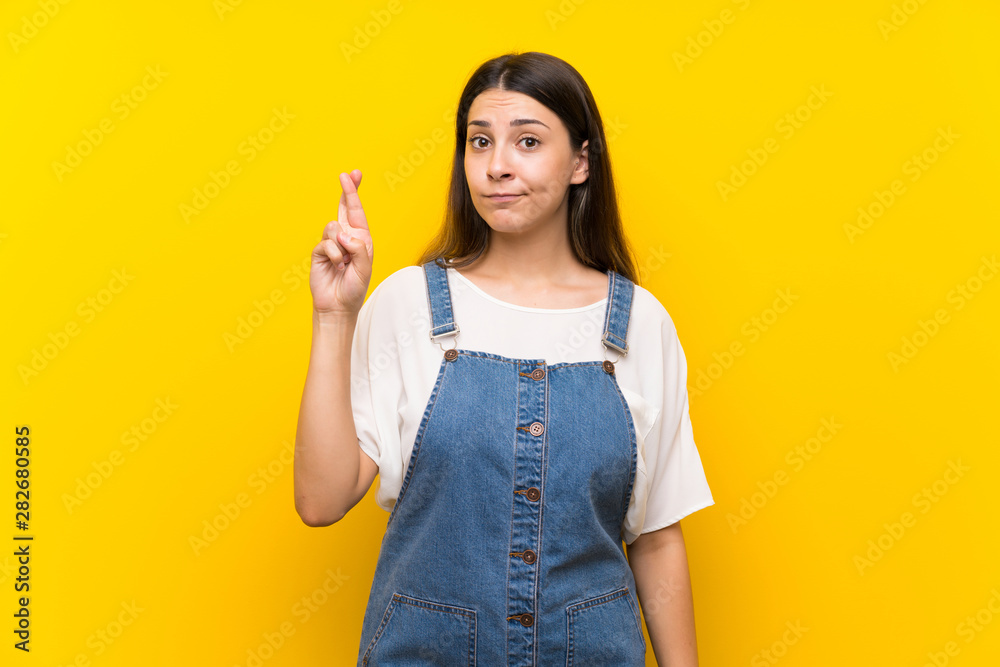 Young woman in dungarees over isolated yellow background with fingers crossing and wishing the best Young woman in dungarees over isolated yellow background with fingers crossing and wishing the best