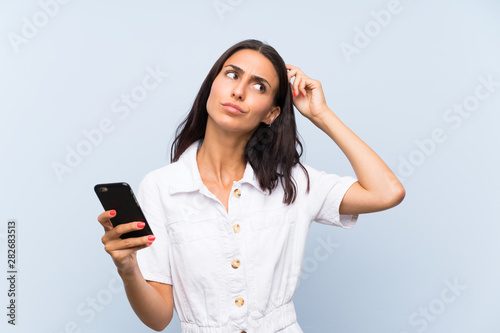 Young woman with a mobile phone over isolated blue wall having doubts and with confuse face expression