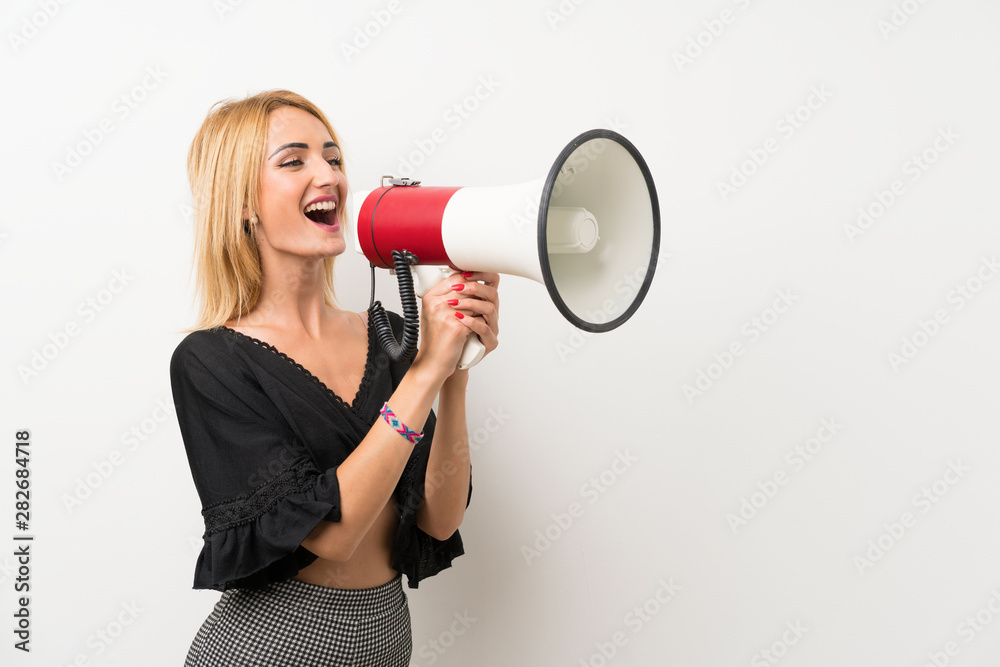 Young blonde woman over isolated white wall shouting through a megaphone Young blonde woman over isolated white wall shouting through a megaphone