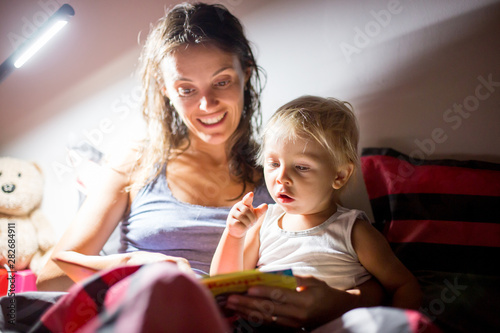 Mother, reading a book to her toddler boy at home at night, night stand lamp turned on
