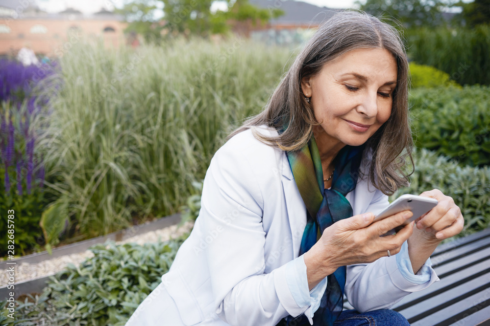 Summertime image of gray haired beautiful retired female sitting on bench with portable electronic gadget, chatting online via messenger. Smiling elegant mature woman using smart phone outdoors - obrazy, fototapety, plakaty 