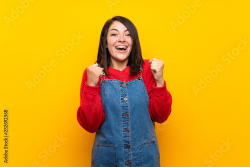 Young Mexican woman with overalls over yellow wall celebrating a victory in winner position