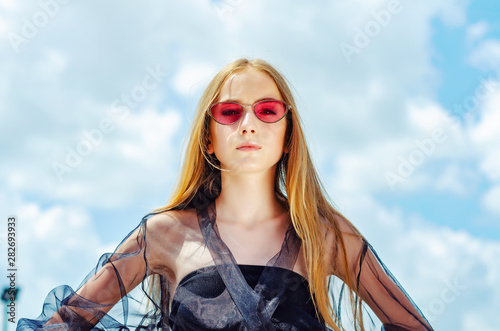 Young girl in red sunglasses posing over sky background, bright sunlight